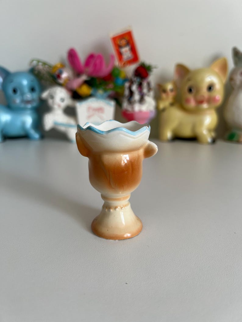 May include: A small, ceramic egg cup with a brown and white glaze. The cup has a scalloped rim and a handle on the side. The cup is sitting on a white surface.