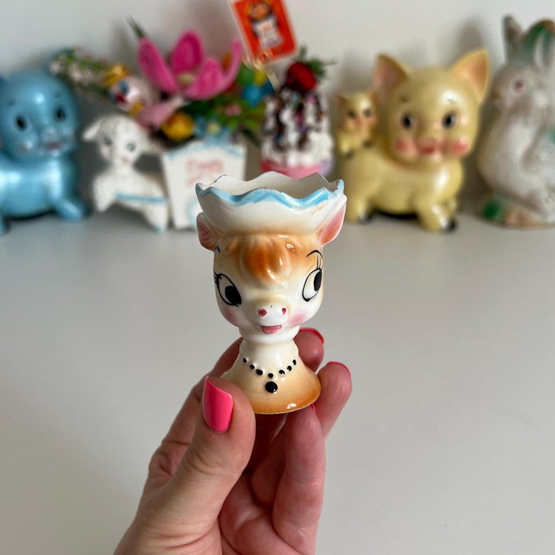 May include: A ceramic cow-shaped egg cup with a blue and white hat, brown eyes, and a black beaded necklace. The egg cup is held in a hand.