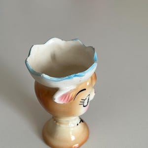 May include: A ceramic egg cup shaped like a cartoon face with blue, pink and brown details. The cup has a white interior and a wide, scalloped rim.