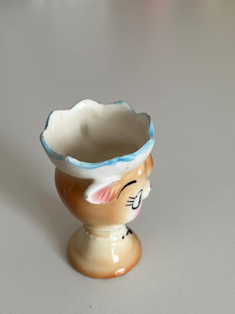 May include: A ceramic egg cup shaped like a cartoon face with blue, pink and brown details. The cup has a white interior and a wide, scalloped rim.