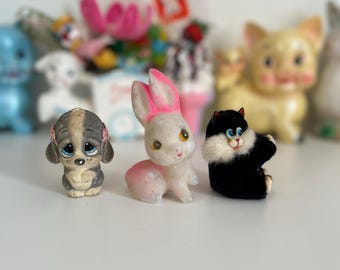 May include: Three small, vintage, toy animals: a gray and white puppy, a white rabbit with pink ears, and a black cat. The toys are made of plastic and have painted details.