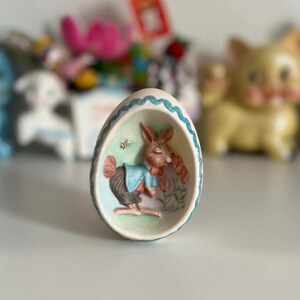 May include: A ceramic Easter egg figurine featuring a 3D image of a bunny wearing a blue shirt and bow tie, smelling a flower. The egg is decorated with a blue and white trim.