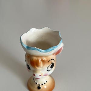 May include: A ceramic egg cup shaped like a cow with a blue and white hat. The cow has brown and white spots and large black eyes.