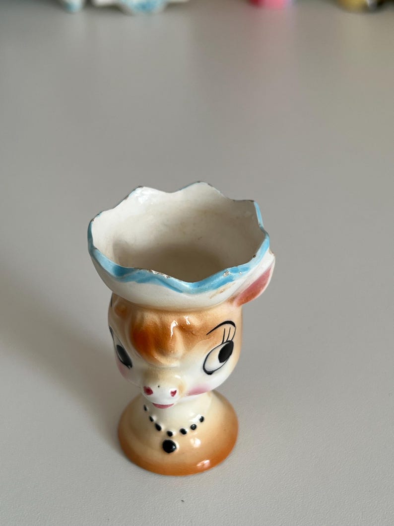 May include: A ceramic egg cup shaped like a cow with a blue and white hat. The cow has brown and white spots and large black eyes.