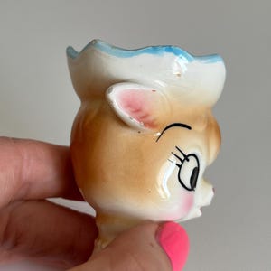 May include: A ceramic egg cup shaped like a cartoon deer with a blue and white hat, pink ears, and brown fur. The deer has a winking eye and a closed mouth.