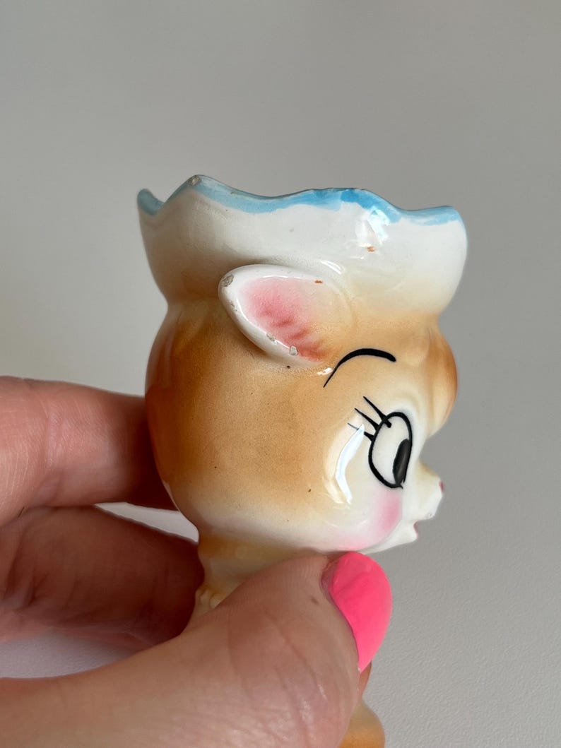 May include: A ceramic egg cup shaped like a cartoon deer with a blue and white hat, pink ears, and brown fur. The deer has a winking eye and a closed mouth.