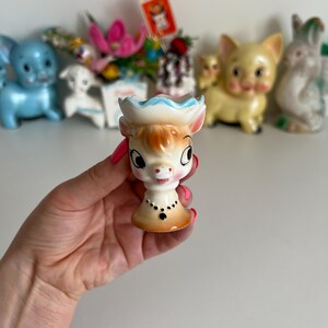 May include: A ceramic egg cup shaped like a cow with a blue and white hat. The cow has brown eyes, a pink nose, and a black necklace.