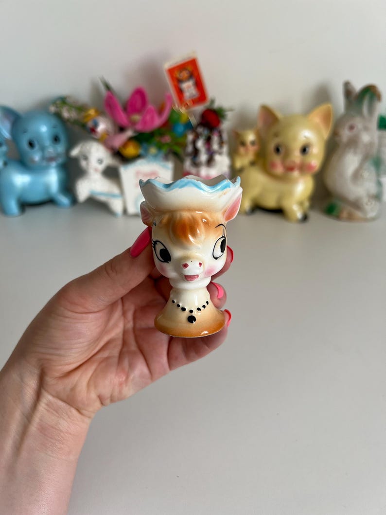 May include: A ceramic egg cup shaped like a cow with a blue and white hat. The cow has brown eyes, a pink nose, and a black necklace.