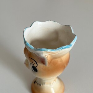 May include: A ceramic egg cup shaped like a cartoon animal with a blue rim and brown and white glaze.