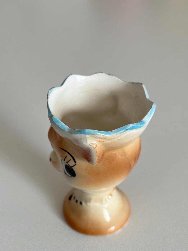 May include: A ceramic egg cup shaped like a cartoon animal with a blue rim and brown and white glaze.