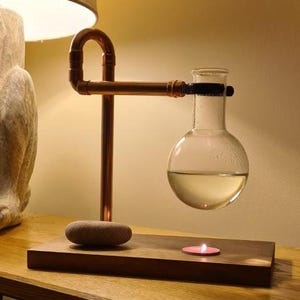May include: A copper pipe diffuser stand with a round glass bottle and a lit candle on a wooden base.