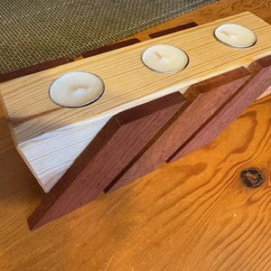 May include: Wooden candle holder with three tealight candles. The holder is made of light-colored wood with dark wood accents. The design is geometric and modern, suitable for home decor.