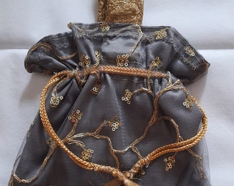 May include: A grey drawstring bag with gold embroidery and tassels. The bag has a gold handle and gold bells on the bottom.