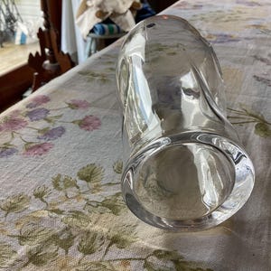 May include: A clear, cylindrical glass vase with a rounded base and a slightly curved body. The vase is empty and sits on a floral patterned tablecloth. The glass reflects light, creating highlights and shadows.