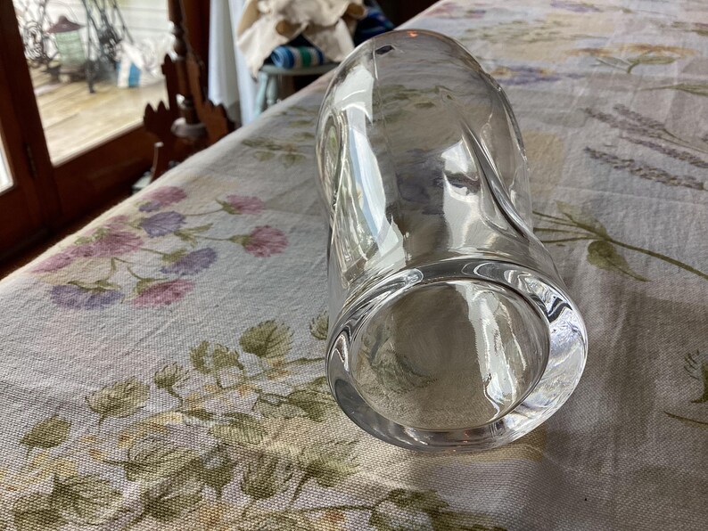 May include: A clear, cylindrical glass vase with a rounded base and a slightly curved body. The vase is empty and sits on a floral patterned tablecloth. The glass reflects light, creating highlights and shadows.