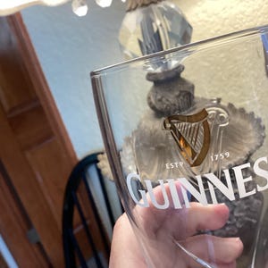 May include: Clear Guinness pint glass with the Guinness logo and harp emblem. The glass has the text "ESTD 1759" and "GUINNESS" in white. The glass is held in front of a blurred background.