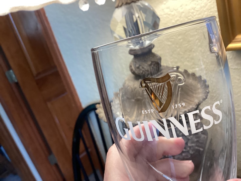 May include: Clear Guinness pint glass with the Guinness logo and harp emblem. The glass has the text "ESTD 1759" and "GUINNESS" in white. The glass is held in front of a blurred background.