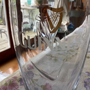 May include: A clear pint glass with the Guinness logo etched on the front. The logo features a harp and the Guinness name in a stylized font. The glass is partially filled and sits on a floral patterned tablecloth.