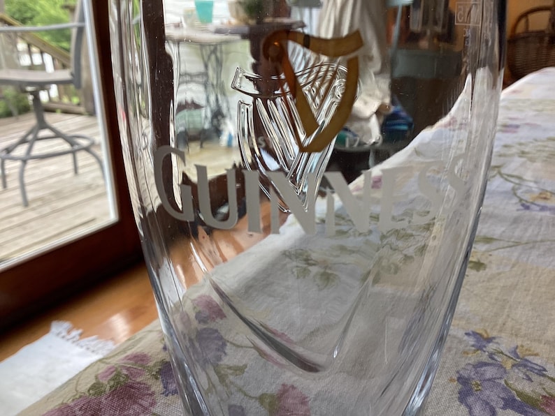 May include: A clear pint glass with the Guinness logo etched on the front. The logo features a harp and the Guinness name in a stylized font. The glass is partially filled and sits on a floral patterned tablecloth.