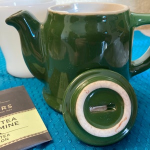 May include: A green ceramic teapot with a white lid and a white rim. The teapot has a handle and a spout. The teapot is sitting on a blue and white patterned surface. The text on the tea bag reads "Taylors Green Tea Jasmine Green Tea Infusion".