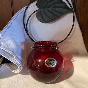 May include: A red glass vase with a black metal leaf design hanging from a ring. The vase has a label that reads "Royal Ruby Anchor Glass".
