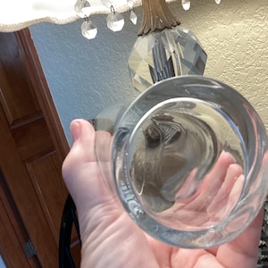 May include: A clear, round glass with a thick base is held up. The glass is empty and reflects the surrounding objects. The glass is part of a lamp with crystal accents.