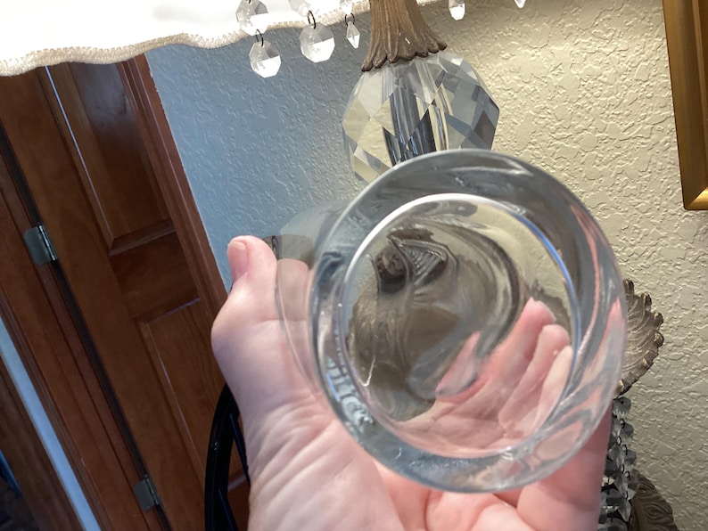May include: A clear, round glass with a thick base is held up. The glass is empty and reflects the surrounding objects. The glass is part of a lamp with crystal accents.