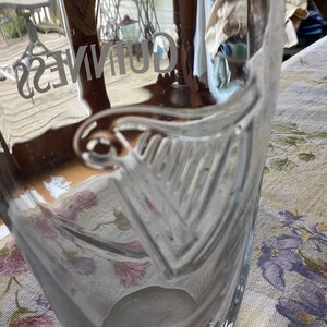 May include: A clear glass Guinness pint glass. The word "GUINNESS" is etched into the glass. The glass features a harp design. The glass is sitting on a floral patterned tablecloth.