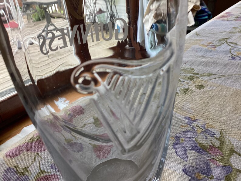 May include: A clear glass Guinness pint glass. The word "GUINNESS" is etched into the glass. The glass features a harp design. The glass is sitting on a floral patterned tablecloth.