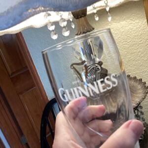 May include: A clear Guinness pint glass with the Guinness logo and text in white. The glass is held in front of a lamp and wooden door. The glass is empty.