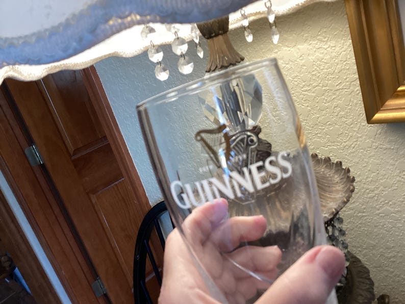 May include: A clear Guinness pint glass with the Guinness logo and text in white. The glass is held in front of a lamp and wooden door. The glass is empty.