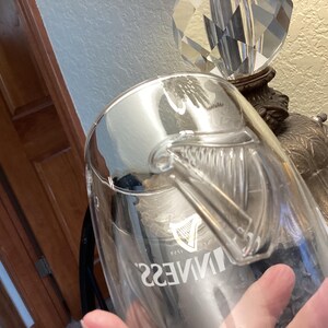 May include: A clear glass Guinness pint glass with a handle. The Guinness logo is visible on the glass. The glass is partially filled and reflects the surrounding environment.