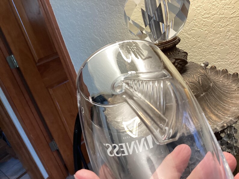 May include: A clear glass Guinness pint glass with a handle. The Guinness logo is visible on the glass. The glass is partially filled and reflects the surrounding environment.