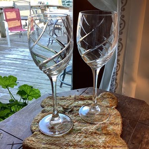 May include: Two clear wine glasses with a crisscross design. The glasses are on a wooden surface with a natural texture. The background includes a deck and outdoor seating.