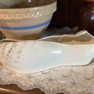 May include: A cream-colored, oblong ceramic planter with a speckled exterior and a light brown interior. The planter sits on a white lace doily, with a large mixing bowl and a dark brown jar in the background. The planter is likely for home decor.