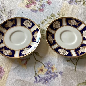 May include: Two white and gold saucers with a navy blue border. The border features gold fish and seashell designs. The saucers are on a floral patterned tablecloth.