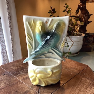 May include: A ceramic wall pocket planter featuring two molded budgerigars in shades of blue and yellow. The birds are perched against a textured background with bamboo-like stalks. The base of the planter is decorated with a raised floral motif.