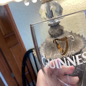 May include: A clear Guinness pint glass with the Guinness logo in white. The logo features a harp and the text "ESTD 1759". The glass is held up in front of a lamp.