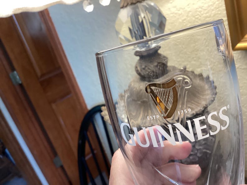 May include: A clear Guinness pint glass with the Guinness logo in white. The logo features a harp and the text "ESTD 1759". The glass is held up in front of a lamp.