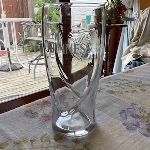 May include: A clear, tall Guinness pint glass with the Guinness logo etched on the front. The glass has a unique, curved design and sits on a floral tablecloth. The background includes a patio and a glimpse of a window.