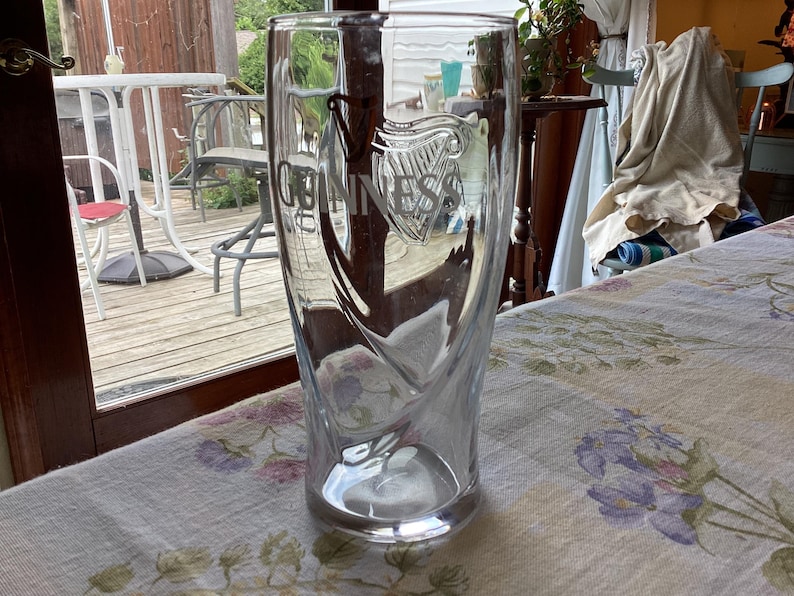 May include: A clear, tall Guinness pint glass with the Guinness logo etched on the front. The glass has a unique, curved design and sits on a floral tablecloth. The background includes a patio and a glimpse of a window.