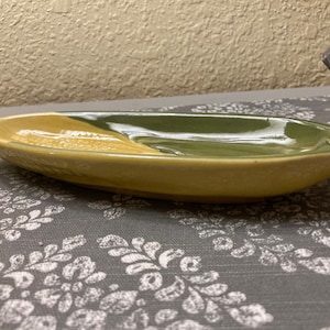 May include: A ceramic serving dish shaped like an ear of corn. The dish is glazed in a light yellow and green color.