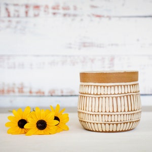 Puede incluir: Un macetero de cerámica blanca con un borde marrón y un patrón geométrico. El macetero está sobre una superficie blanca con tres girasoles amarillos delante.