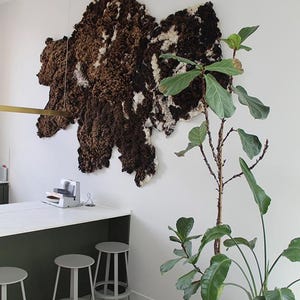 Op de afbeelding: Een witte muur met een grote bruine en witte wanddecoratie van wol. Een groene plant met grote bladeren staat in een pot in de hoek van de kamer. Er staan drie grijze krukken voor een wit aanrecht.