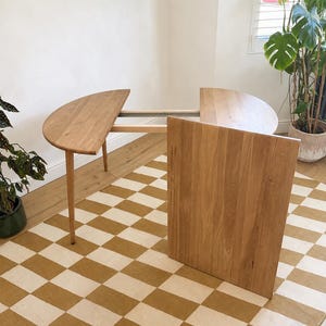 May include: A light brown wooden dining table with a semi-circular end and an extendable mechanism. The table features a rectangular leaf panel positioned vertically in the front, revealing the internal metal and wood support structure.