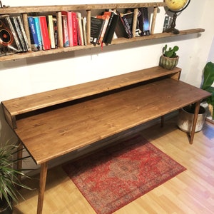 May include: A long wooden desk with a shelf above it. The desk has a natural wood finish and is supported by two legs. The shelf is made of reclaimed wood and has a ladder design. There are books on the shelf and a globe on the wall behind the desk.