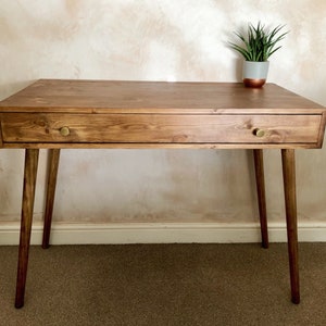 May include: A wooden desk with a single drawer and tapered legs. The desk has a dark brown stain and brass drawer pulls. The desk is sitting on a brown carpet in front of a light pink wall.