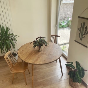 May include: A round wooden dining table with a natural wood finish and three legs. The table is set in a room with light wood floors and a window with a view of a garden.