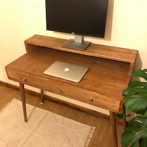 May include: A wooden desk with a light brown finish and a drawer. The desk has a shelf above the drawer for a monitor. The desk has a laptop on top of the drawer and a monitor on the shelf. The desk has two legs on each side. The desk is sitting on a light brown rug with a geometric pattern. A green plant is in the background.