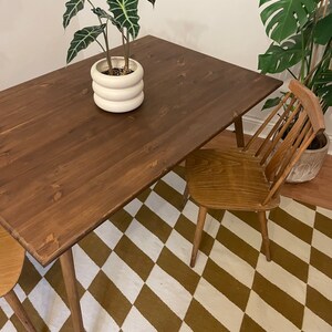 May include: A dark brown wooden dining table with a white ceramic planter and a green plant on top. The table has four legs and is sitting on a brown and white checkered rug. A wooden chair is pulled up to the table.